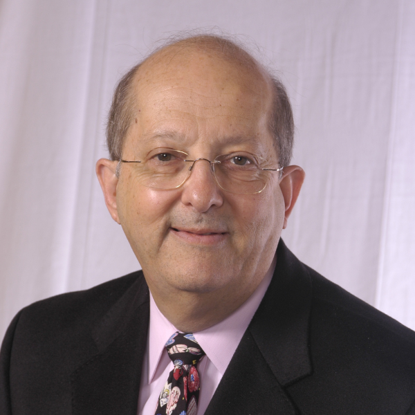 A man wearing a black suit, pink shirt, patterned tie, and glasses smiles at the camera.