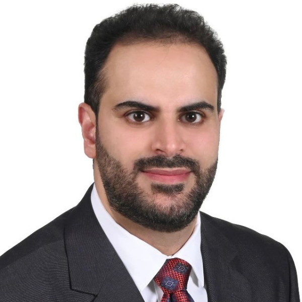 A man with dark hair, a beard, and wearing a black suit and red tie.