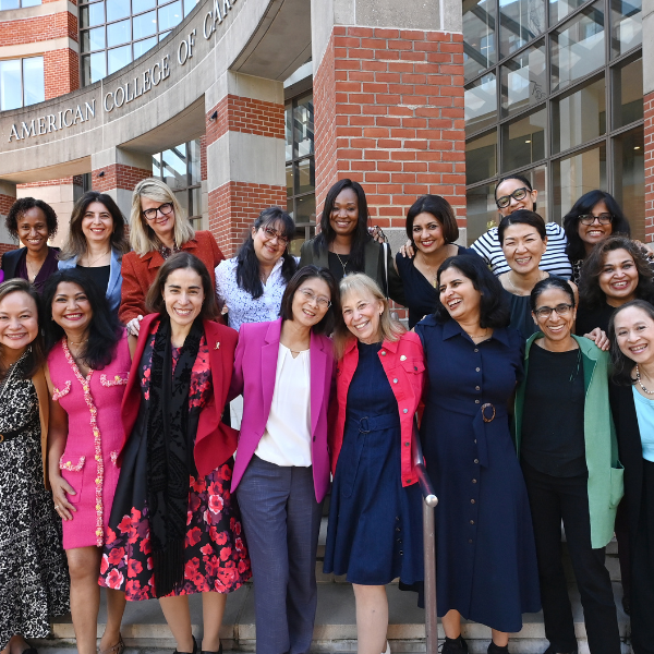 A group of women stand outside a brick building. They are smiling at the camera and have their arms wrapped around each other.