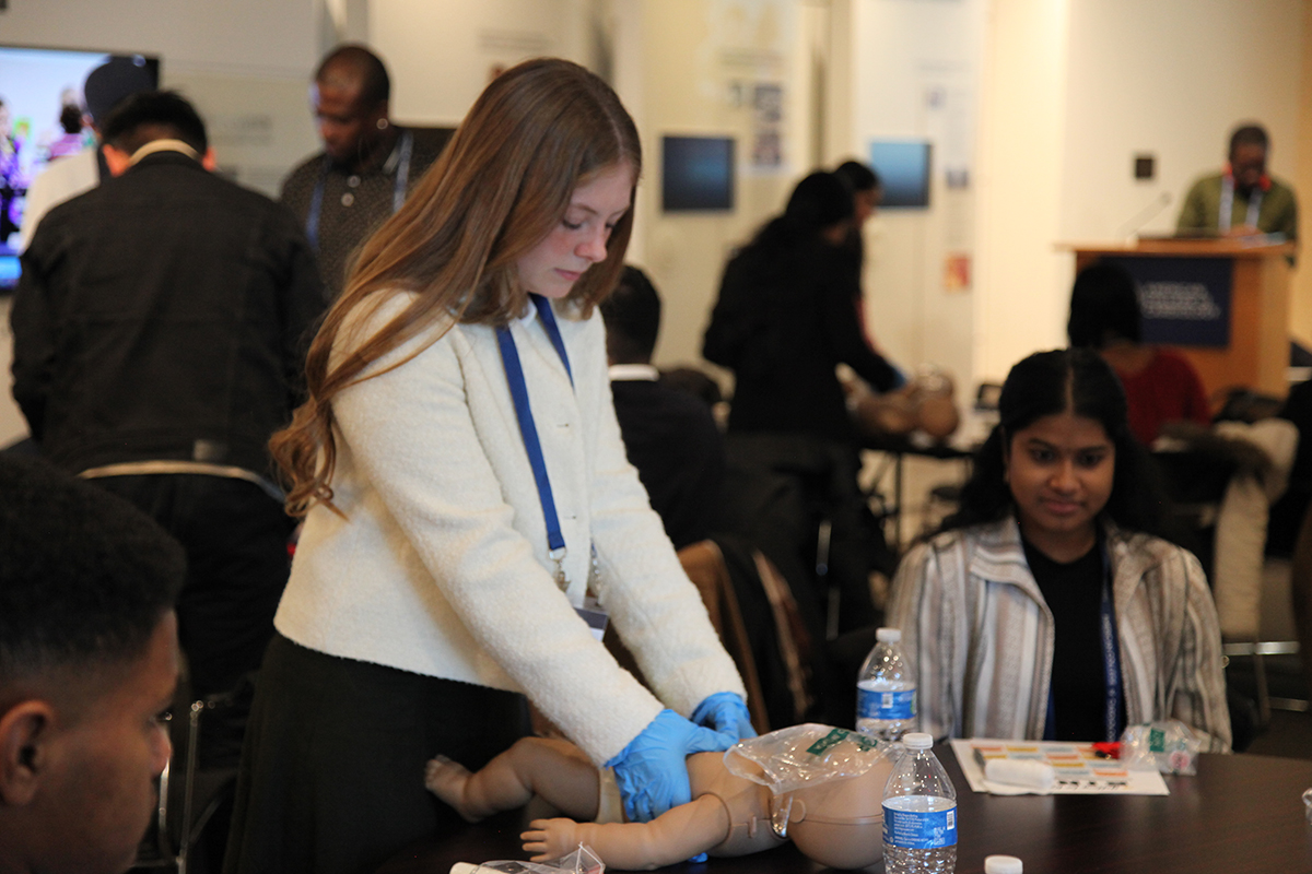 A room full of students learning CPR techniques on infant dolls. The focus is on a woman in a white blazer wearing blue medical gloves with her hands wrapped around the doll as it lays on a table.