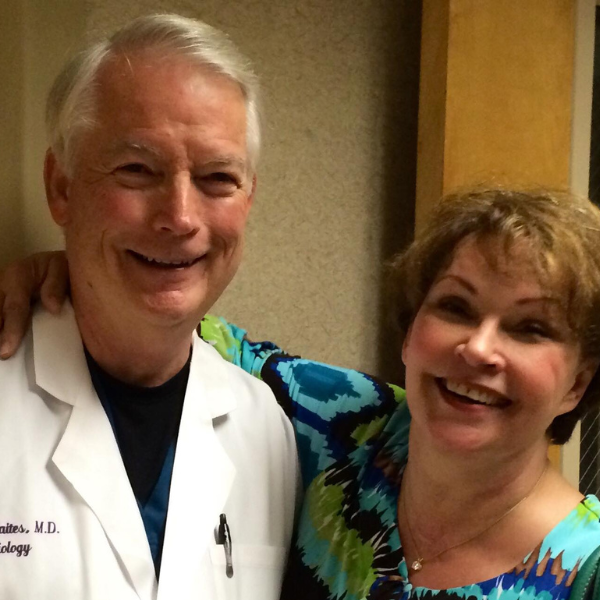 A smiling man in a white lab coat looks at the camera. A woman in a blue floral dress has her arm over the man's shoulder as she also smiles at the camera.