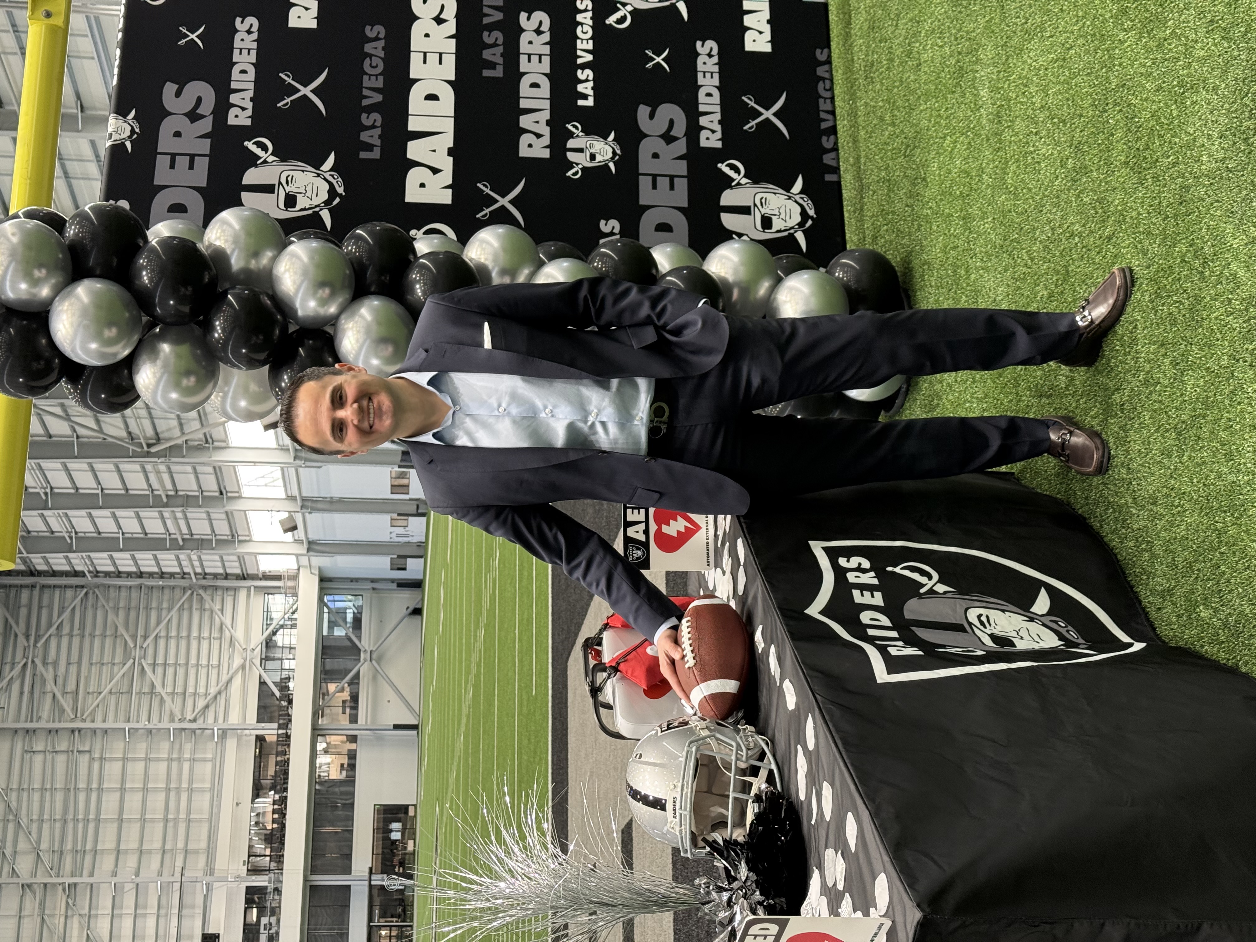 Person standing beside a display table with Las Vegas Raiders branding, football equipment, and an AED device inside an indoor practice field.