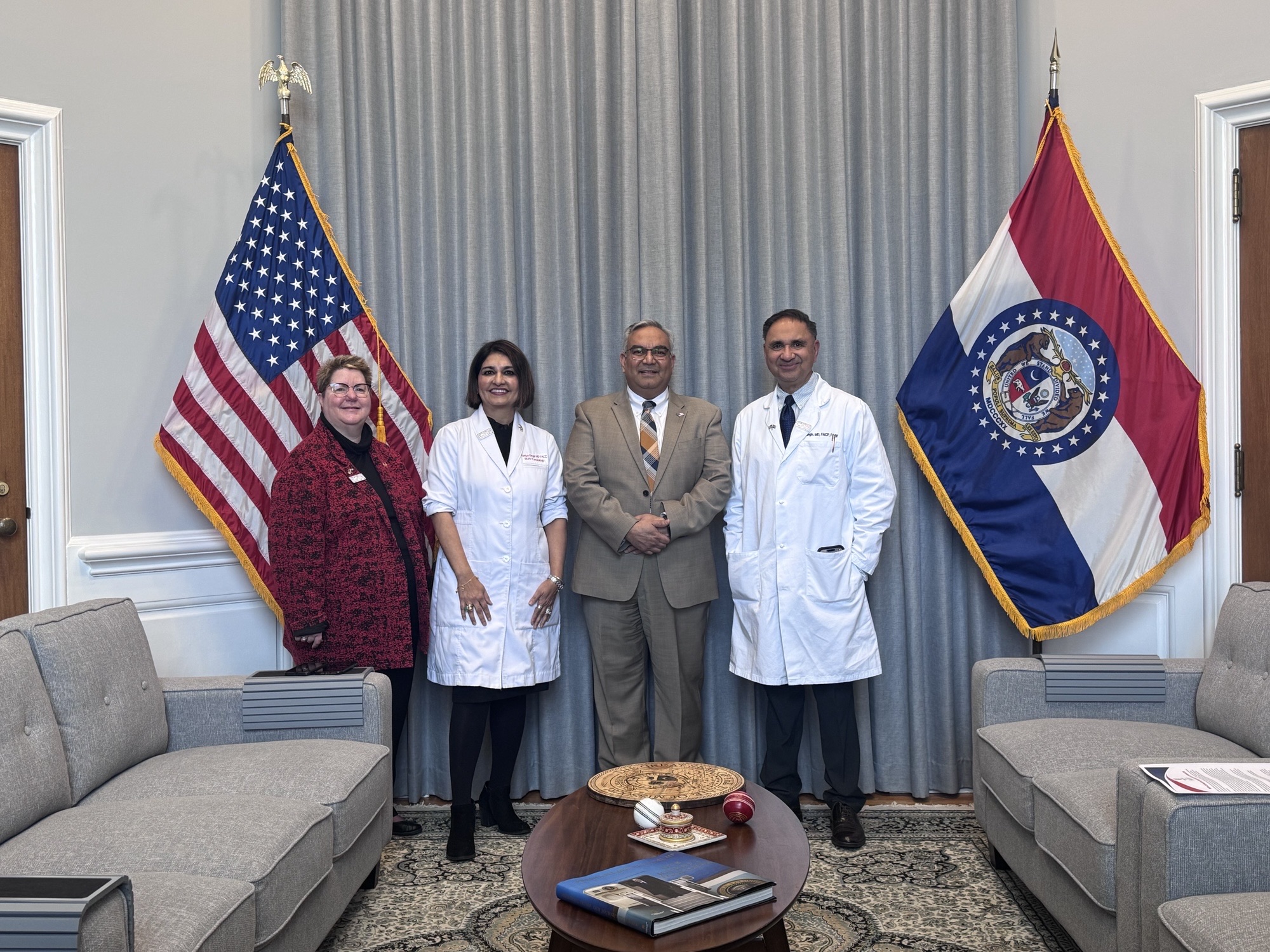 Several individuals, including two people in white coats, stand with a government official in an office featuring the U.S. flag and a state flag