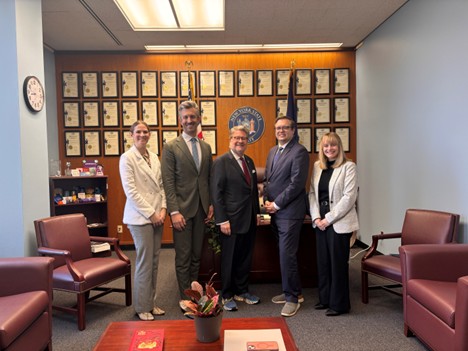 Group of people in business attire standing together in a legislative office with framed certificates covering the back wall